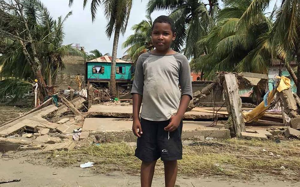 Ein Junge steht vor zerstörten Gebäuden und umgekippten Palmen. 