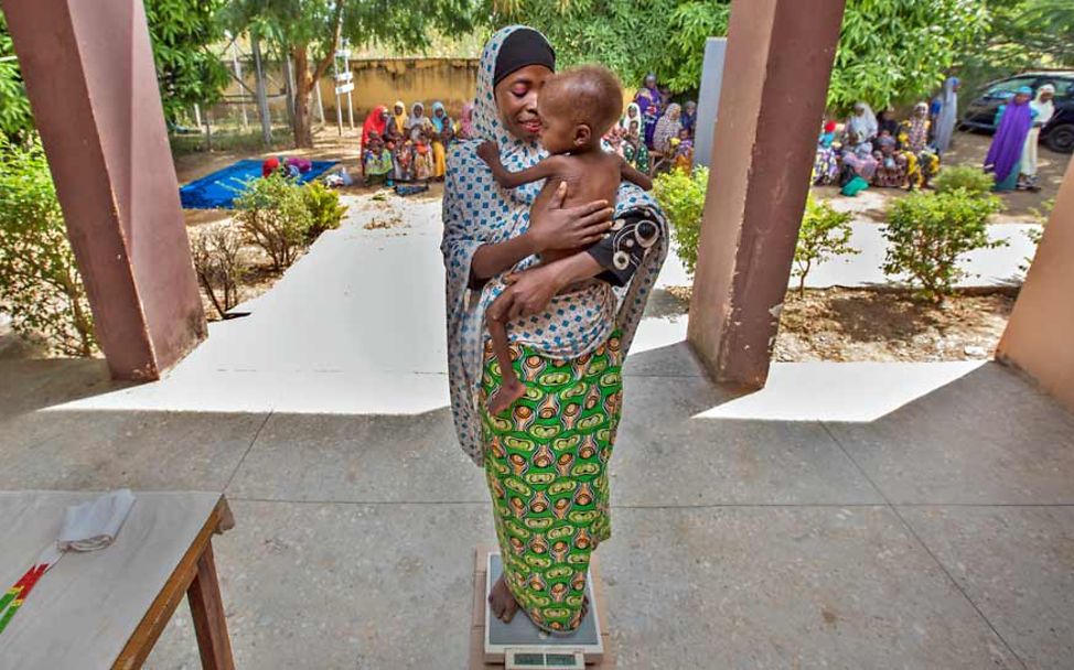 Eine Mutter steht mit ihrem Baby auf dem Arm in einem überdachten Außenbereich auf einer Personen-Waage.