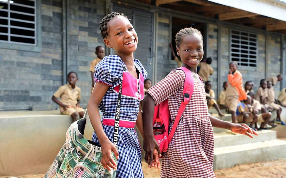 Zwei Mädchen gehen Hand in Hand und lachend vor ihrer aus Plastikziegeln gebauten Schule vorbei.