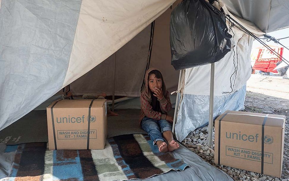 Ein Junge sitzt barfuß in einem Nothilfe-Zelt, neben ihm stehen UNICEF-Kartons mit Hilfsgütern.