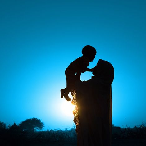 Eine Mutter in einem afrikanischen Dorf hält ihr Kleinkind über sich in den Himmel. 