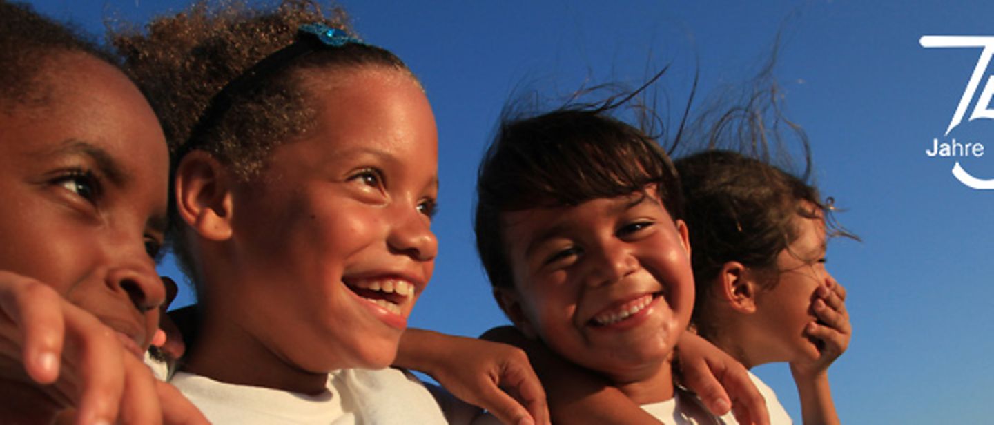Vier Mädchen in Aruba stehen lachend Arm in Arm vor einem blauen Himmel. 