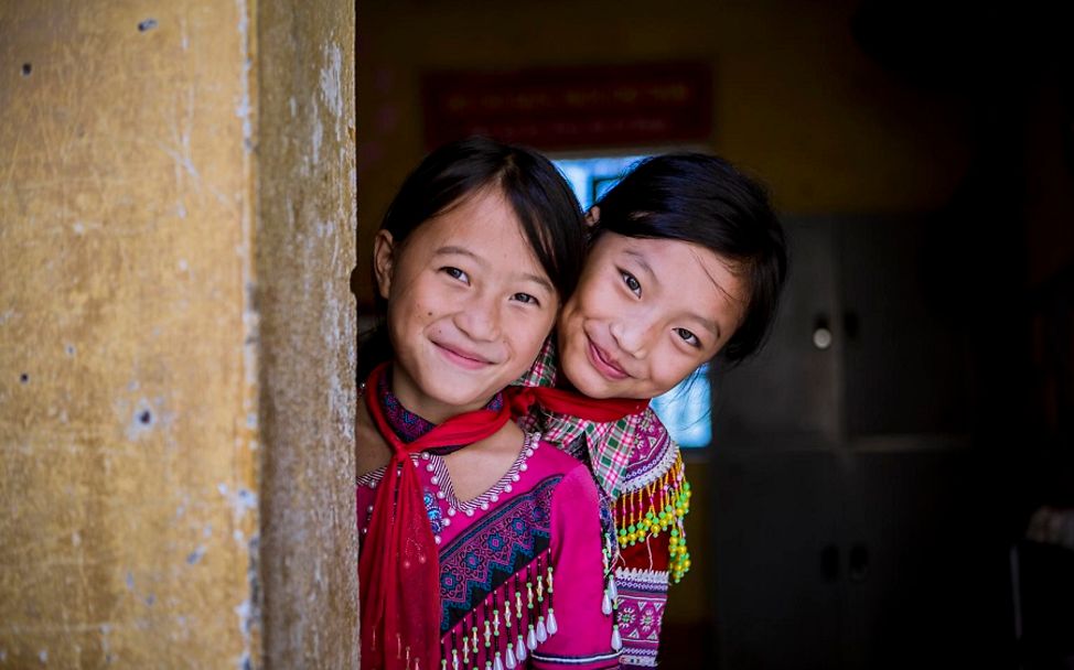 Viertnam: Zwei Mädchen gucken hinter einer Mauer hervor und lächeln.