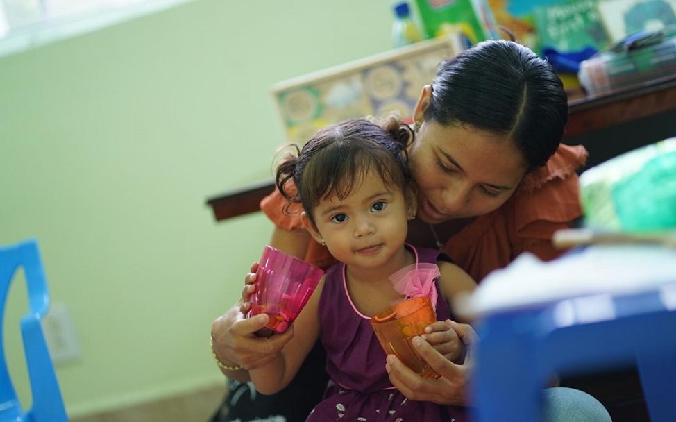 Belize: Tochter sitzt auf dem Schoss ihrer Mutter bei einer Routineuntersuchung beim Arzt