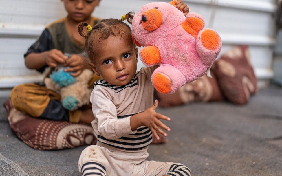 Jemen: Mädchen in Notunterkunft hält Teddy hoch.