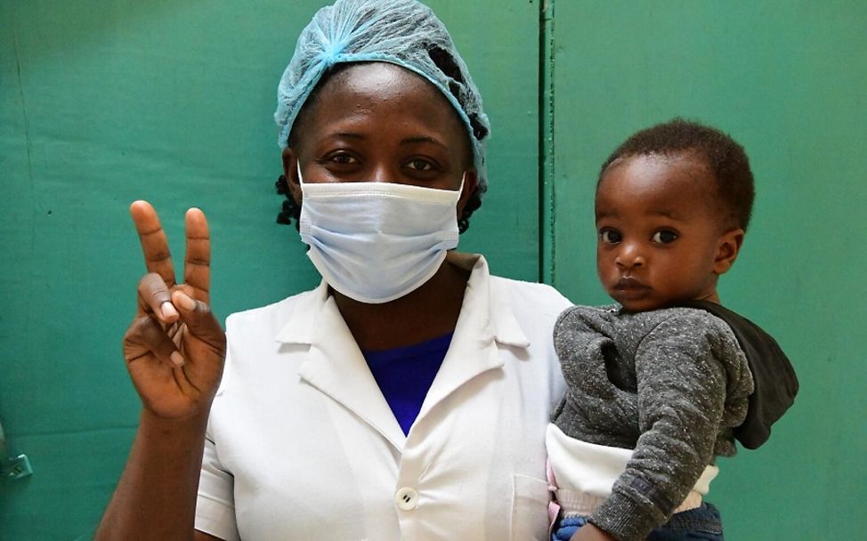 Kamerun: Geimpfte Gesundheitshelferin mit Baby auf dem Arm zeigt ein V für Vaccinated.