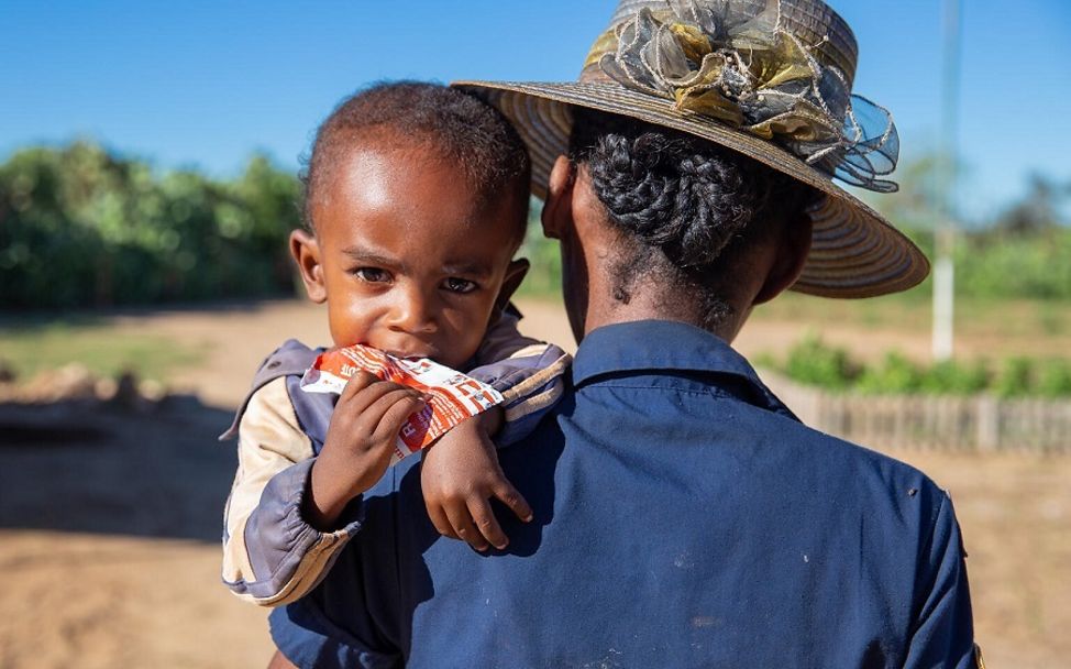 Madagaskar: Kind auf dem Arm der Mutter hält Erdnusspaste in der Hand 