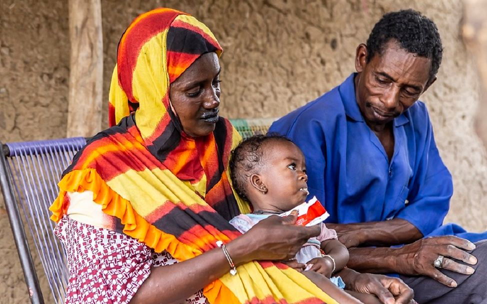 Mali: Mutter und Vater haben ihr Kind auf dem Schoß und füttern es mit therapeutischer Nahrung.
