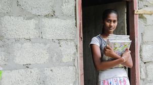 Dilrukshi möchte Lehrerin werden. Sie steht mit Schulbüchern in der Hand vor ihrer Haustür.