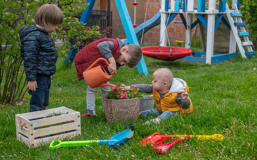 Serbien: Drei Kinder spielen im Garten und gießen eine Blume mit einer Gießkanne.