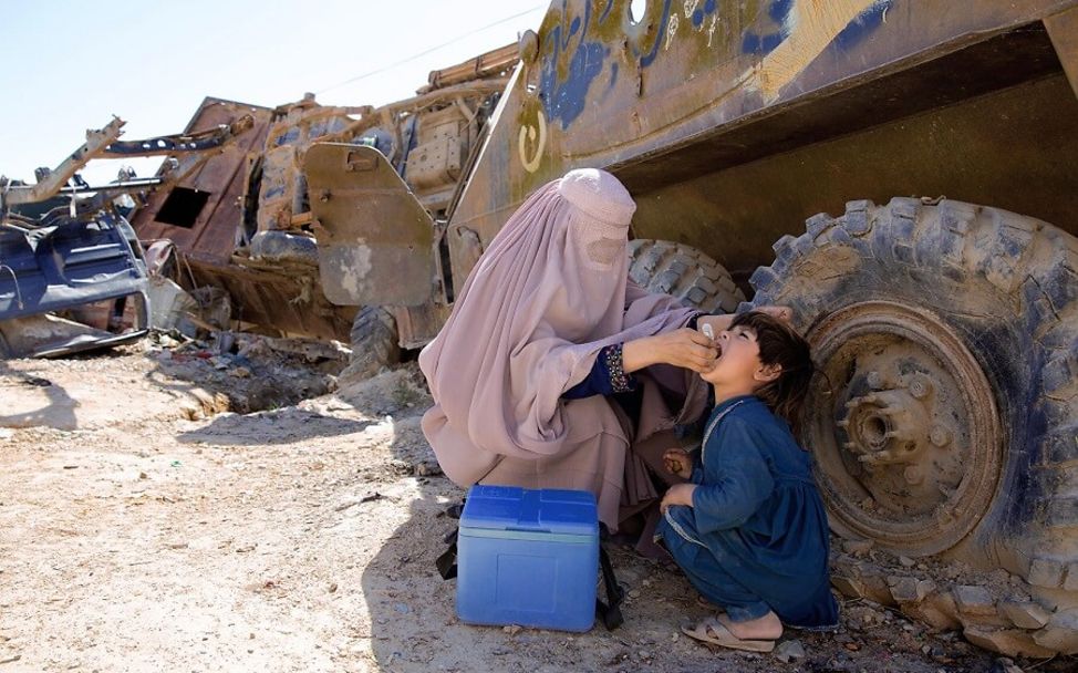 Afghanistan: Eine Helferin gibt einem Jungen eine Schluckimpfung zwischen zerstörtem Kriegsgerät