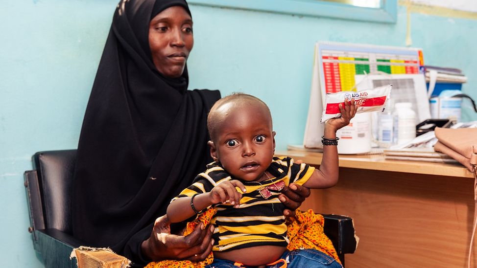 Krankenhaus in Kenia: Mutter hat Kind auf dem Schoß, das ein Päckchen Erdnusspaste in der Hand hält.