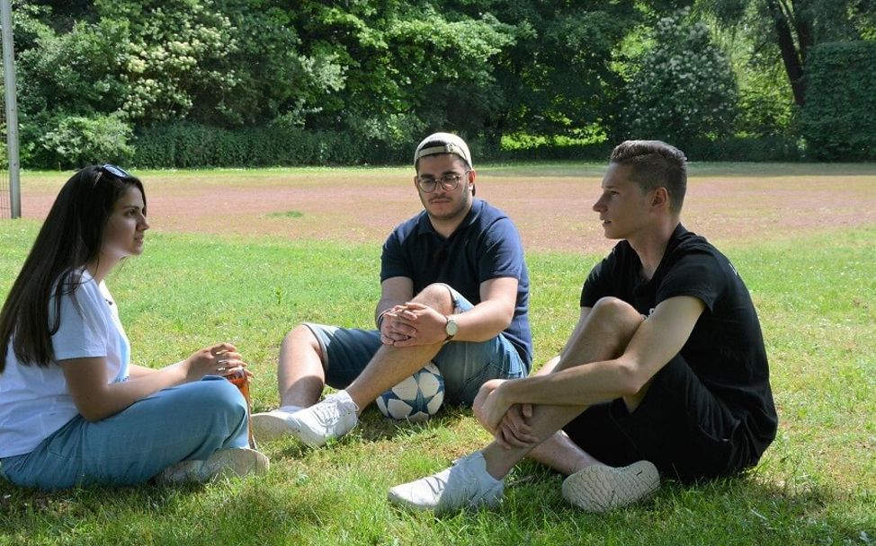 Julian Draxler sitzt mit Shaza und Berkay auf der Wiese eines Bolzplatzes.
