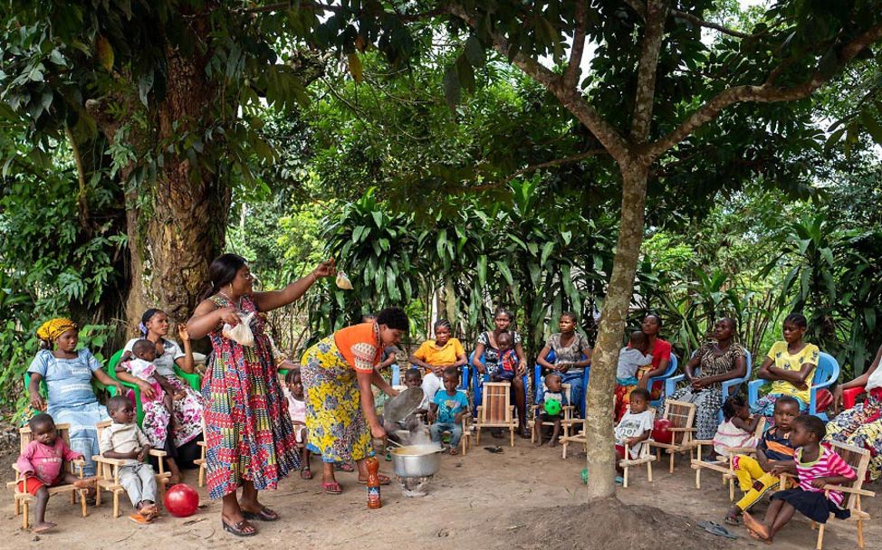 Kongo: Frauen und Kinder sitzen um eine Frau herum, die an einem Kochtopf Kochen demonstriert.