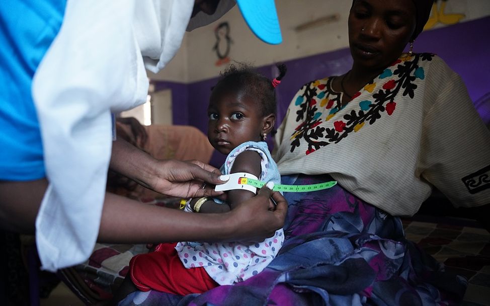 Sudan: Einem kleinen Mädchen, das auf dem Schoß der Mutter sitzt, wird ein Messband um den Arm gelegt.
